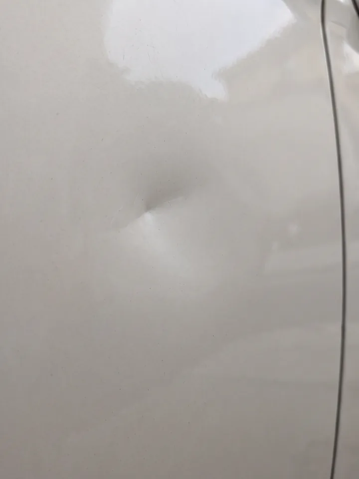 Avant réparation : bosse de coup de portière sur panneau blanc, déformation visible dans le reflet
