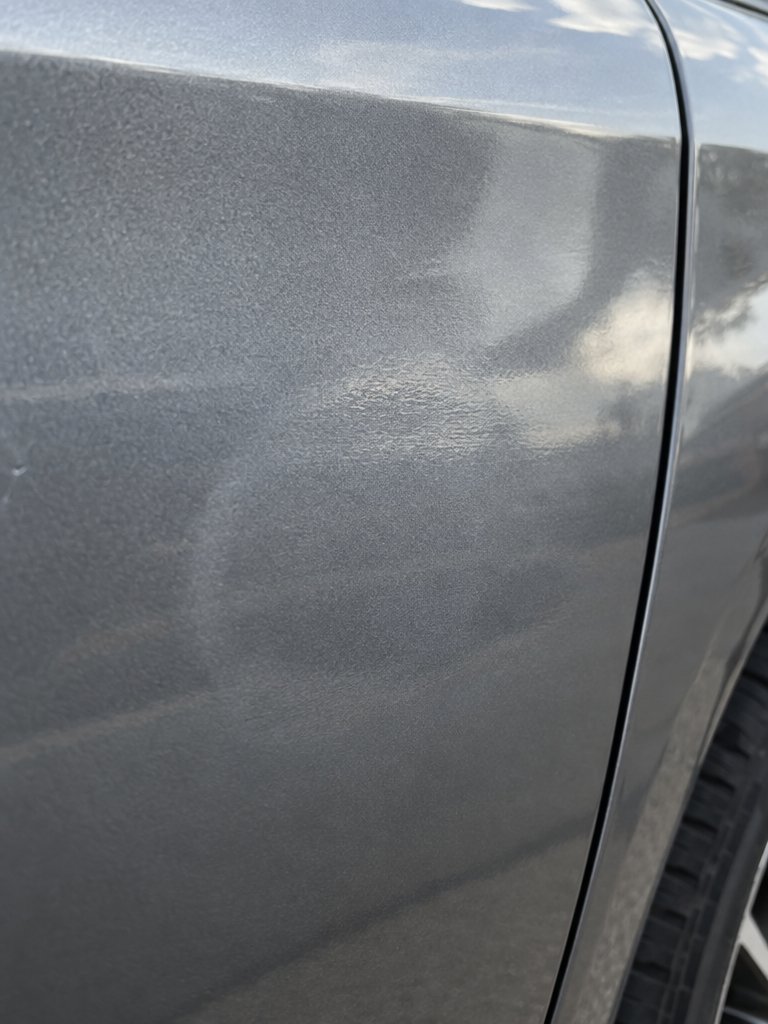 Gros plan sur une portière de voiture grise métallisée : la marque de choc se révèle dans le reflet déformé de la peinture brillante